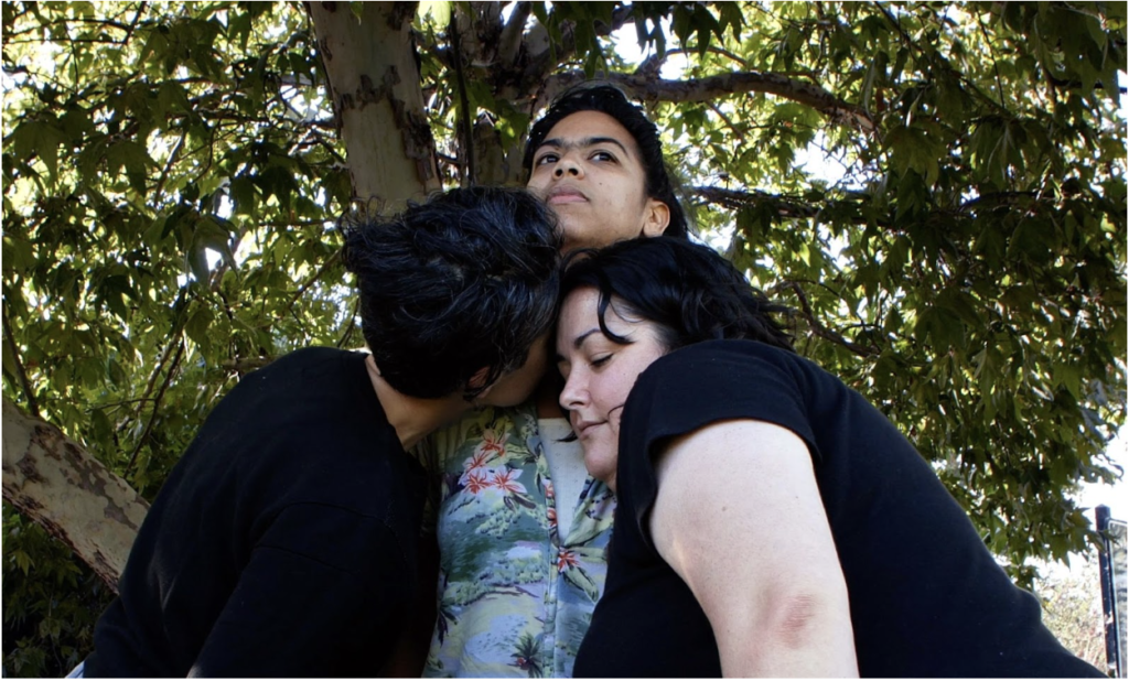 Photo of three people under a tree. Two people have their face pressed close to the middle person's chest. A mental person is standing tall gazing forward.