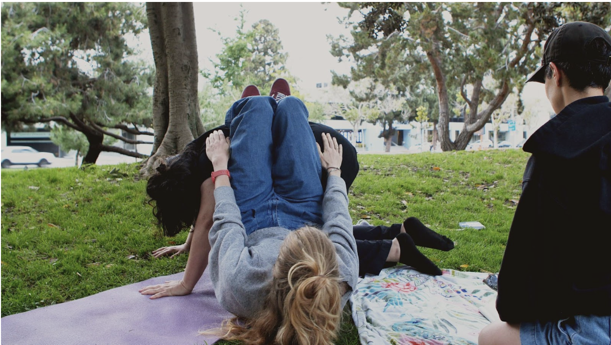 A photo of three people outdoors in a grassy park. One person sits apart, the other two are in contact on a yoga mat.