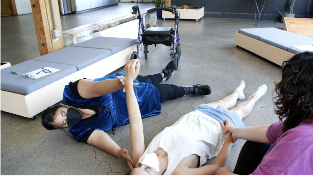 Photo of three people on the ground. The person in the middle is having their arms lifted by the two people on either side. There is an assistive walking device, and benches nearby.